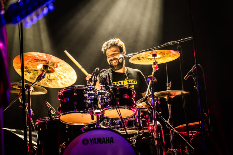 Een drummer speelt enthousiast op een set drums onder achtergrondverlichting in een optreden.