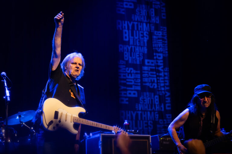 Een muzikant met een gitaar steekt zijn arm omhoog tijdens een optreden op een sfeervol verlicht podium.