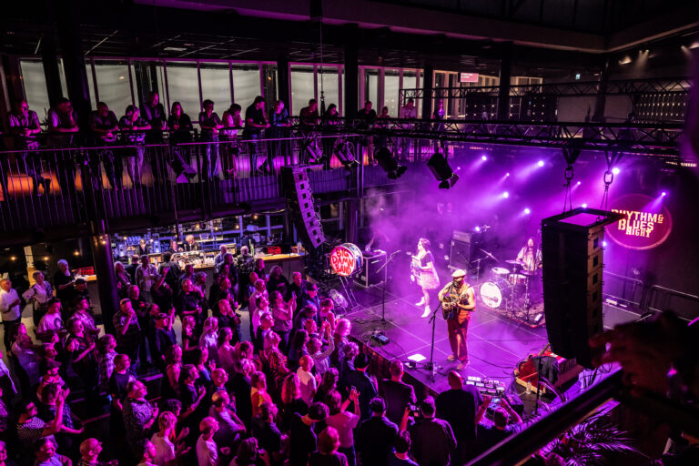 Een levendig concert met een zingende artiest op het podium, omringd door een enthousiaste menigte en kleurrijke verlichting.