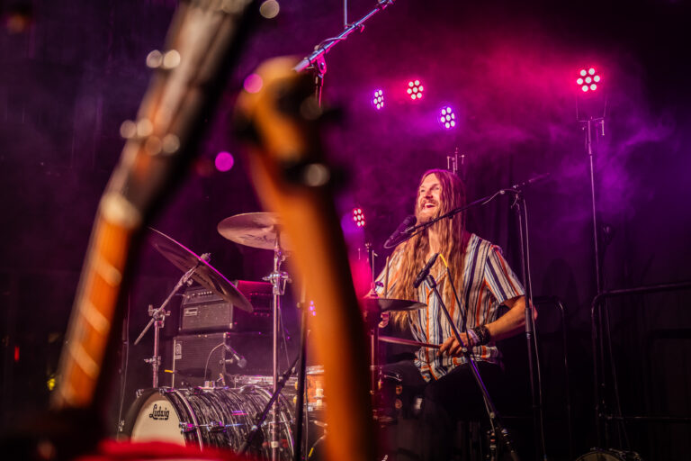 Een drummer speelt op een podium, omgeven door sfeerverlichting en een mistige atmosfeer.