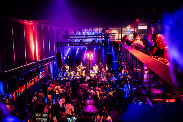 Een drukke concertzaal met een band die op het podium speelt en kleurrijke verlichting.