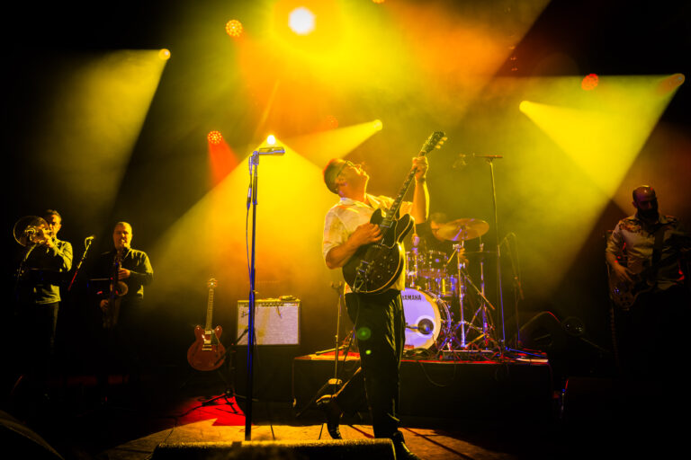 Een muzikant speelt gitaar op een podium met felgekleurde lichtstralen en een drummer op de achtergrond.