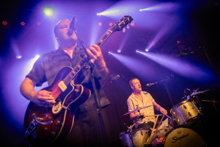 Twee muzikanten optreden op een podium, met een gitarist aan de voorkant en een drummer daarachter, verlicht door opvallende lichten.