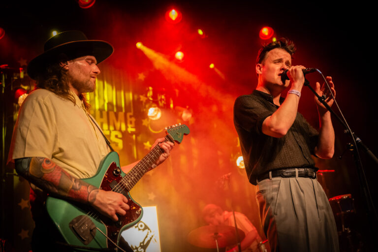 Twee muzikanten staan op een sfeervol verlichte scène, één met een gitaar en de ander zingt in een microfoon.