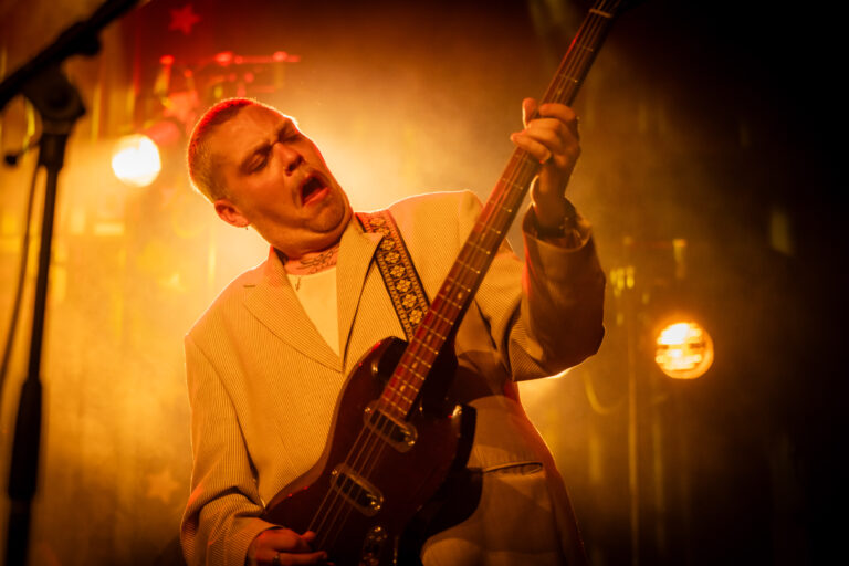 Een muzikant speelt enthousiast gitaar op een podium, omringd door sfeervolle lichten.