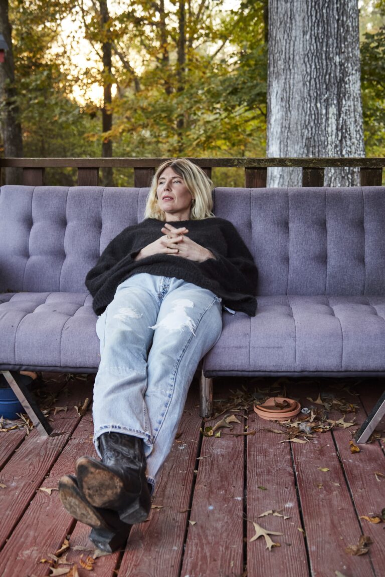 Een vrouw ligt ontspannen op een paarse bank op een houten terras, omgeven door herfstbladeren en bomen, gekleed in een zwarte trui en gescheurde jeans, met haar handen gevouwen over haar buik.