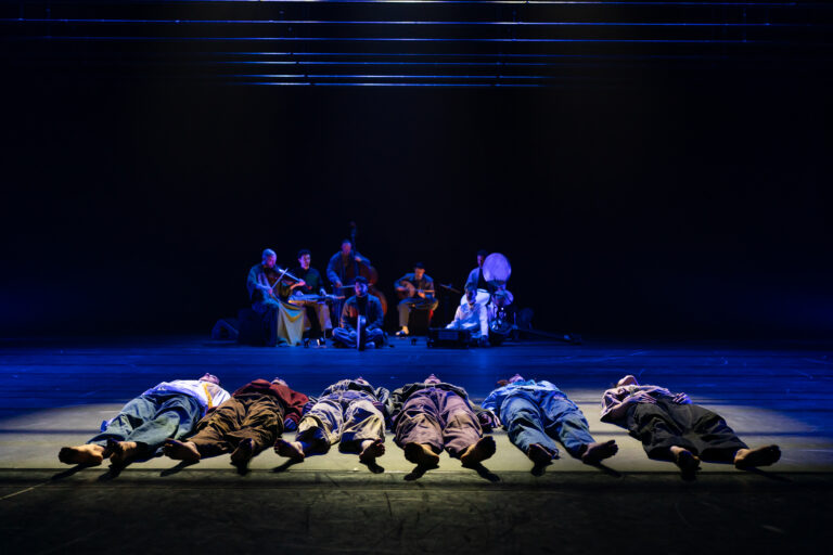 Een groep performers ligt op de vloer van een podium, terwijl een muzikale groep op de achtergrond speelt.
