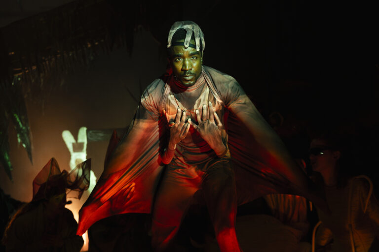 Een performer op een donker podium met een lange rode cape die zich uitspreidt over de scène.