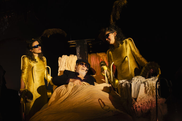 Een persoon ligt in een bed op een toneel, omringd door meerdere figuren in gele kostuums.