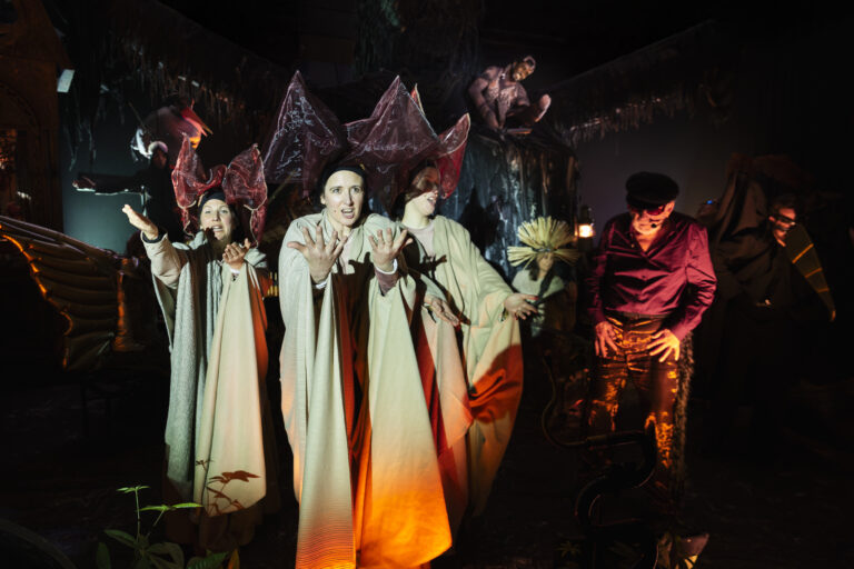 Een groep acteurs in witte gewaden staat op een donker toneel, verlicht door warme oranje gloed.