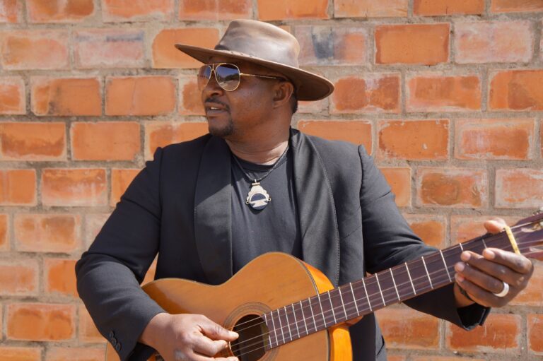 Man met hoed, zonnebril en zwarte kleding kijkt naar links en speelt gitaar voor oranje stenen muur.