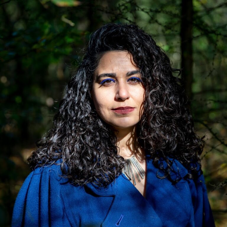 Vrouw met krullend haar en blauwe make-up, staande in een bos met natuurlijk licht.