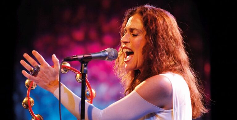 Een vrouwelijke zangeres performt met een microfoon en een tamboerijn op een kleurrijk verlichte achtergrond.