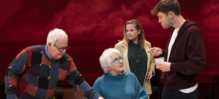 De acteurs staan met z'n allen rondom de tafel. Er is een rode achtergrond.