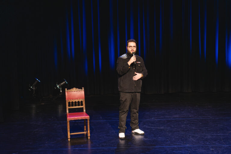 Een man staat op een podium en spreekt in een microfoon, naast een houten stoel tegen een donkerblauwe gordijnachtergrond.