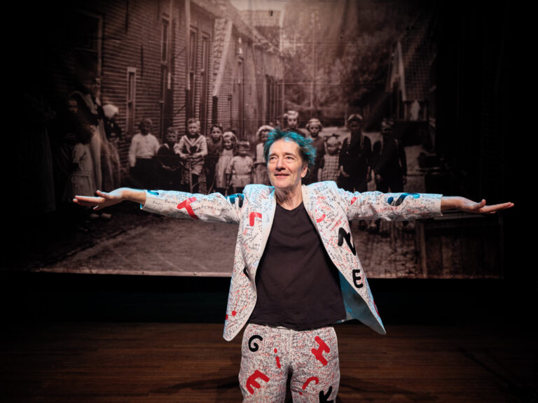 Een persoon met blauwe haren staat met uitgestrekte armen in een opvallend gekleed kostuum voor een zwart-wit achtergrond van een straat vol mensen.