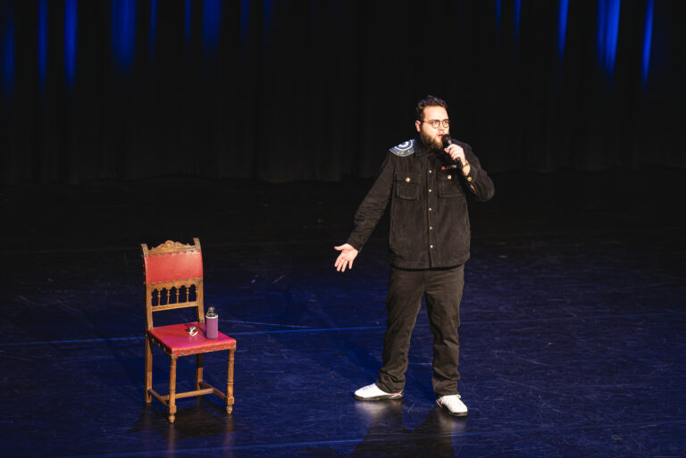 Een comedian staat op een podium naast een lege stoel en spreekt in een microfoon.
