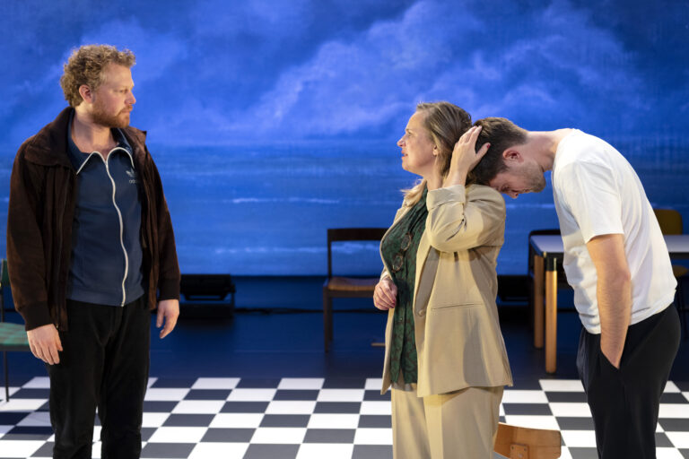 Twee acteurs in een theaterproductie, met de ene acteur staand en de andere gebogen, terwijl een derde acteur in het midden staat.