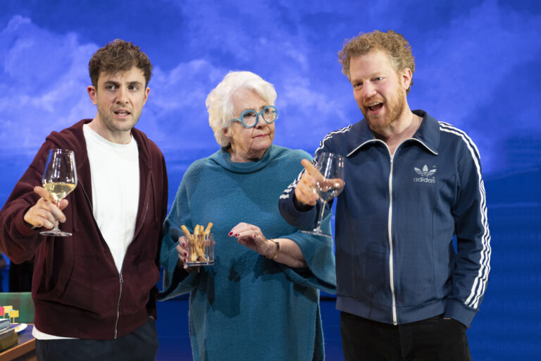 Drie mensen staan samen met glazen in de hand, kijkend naar de camera met een kleurrijke achtergrond.