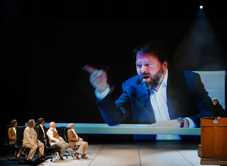 Een man met baard en pak wijst terwijl hij aan een lange tafel zit tijdens een paneldiscussie, met andere panelleden op de achtergrond.