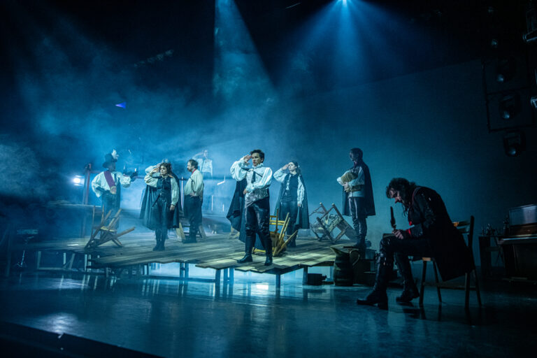 Een groep artiesten staat op een verlicht podium met blauwe lichten tijdens een optreden.