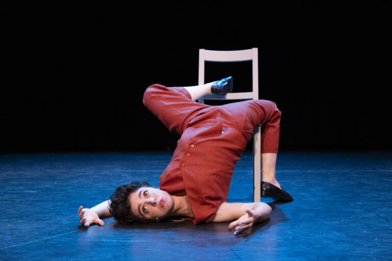 Een performer, Selma Visscher, in een rood kostuum ligt op de grond in een kunstzinnige pose, met één been op een witte stoel en een intense blik gericht naar de camera, op een donkere toneelachtergrond.