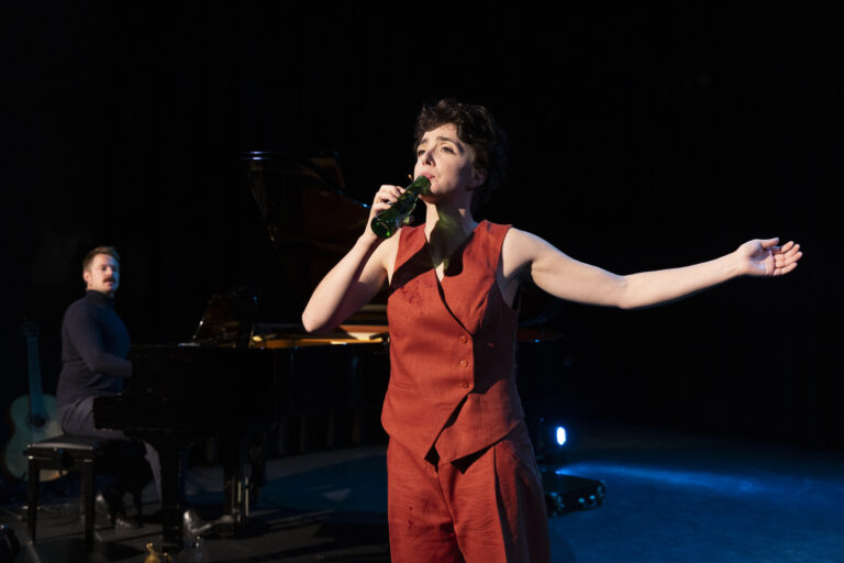Selma Visscher staat op een donkere toneelachtergrond, gekleed in een rood ensemble, terwijl ze uit een flesje drinkt met haar andere arm wijd uitgestrekt; op de achtergrond zit een pianist achter een vleugel.