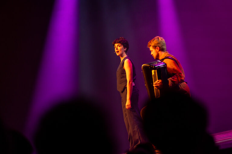 Selma Visscher staat op het podium, gekleed in een donker ensemble, terwijl ze zingt onder paarse podiumverlichting; naast haar speelt een muzikant accordeon.