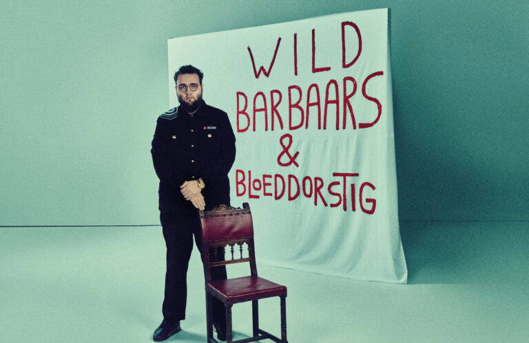 Sezgin staan achter een stoel met een groot spandoek met de titel van de voorstelling achter hem