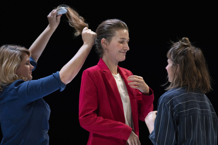 Drie vrouwen zijn actief bezig met het stylen van het haar van een vrouw in een rode blazer op een podium met een donkere achtergrond.