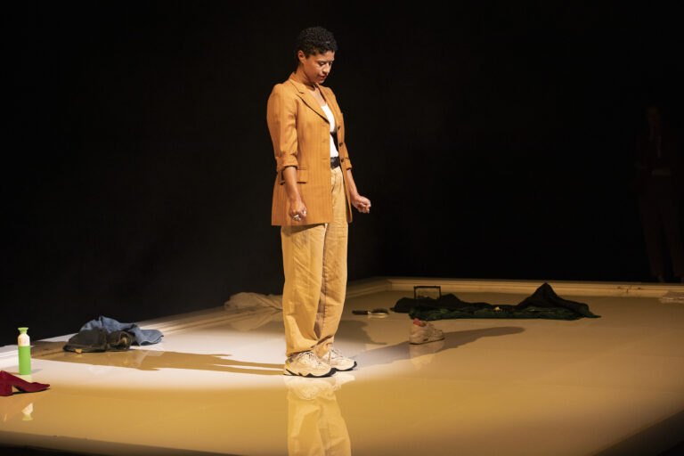 Een vrouw staat op een glanzend podium te midden van kledingstukken en andere voorwerpen in een donkere omgeving.
