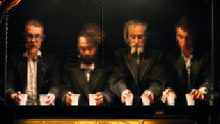 Vier mannen die kijken naar hun handen waarin ze twee bekertjes vast hebben. Ze zijn verkleed als een soort goochelaar