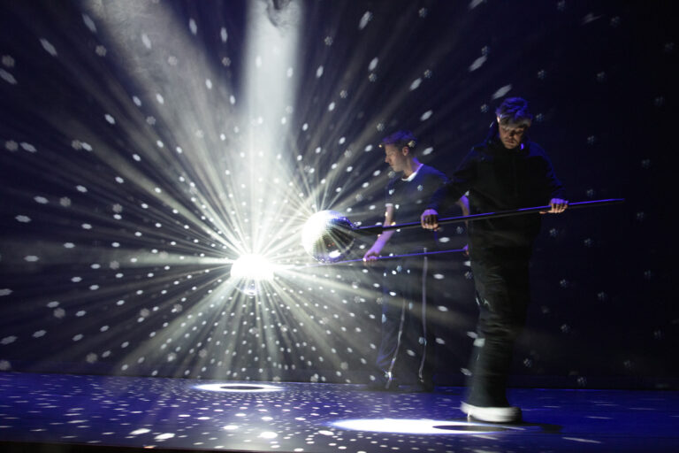 Twee performers staan op een podium met een opvallende lichtshow en stralen licht in verschillende richtingen.