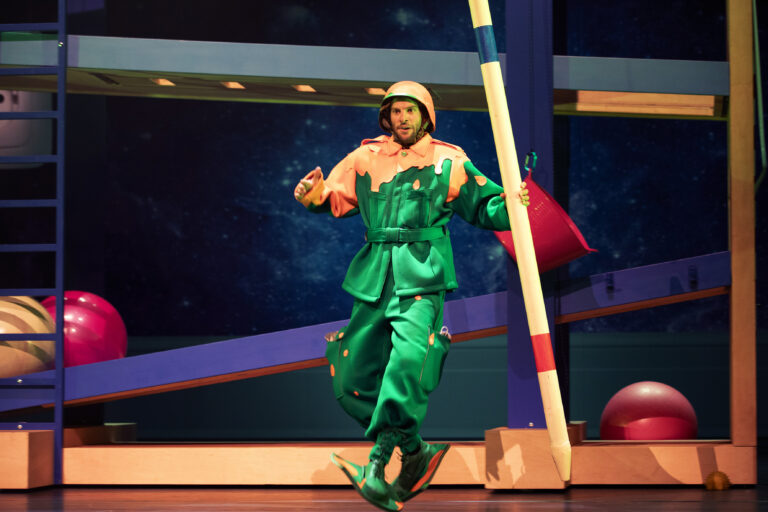 Een performer in kleurrijke kleding staat op een evenwichtsbalk met een speelse achtergrond van grote ballen en een littekenstructuur.