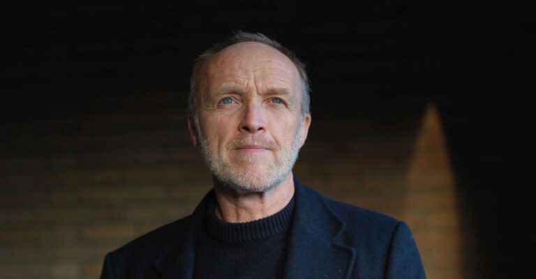 Stef Bos staat in een donkere ruimte. Hij kijkt voor zich uit.
