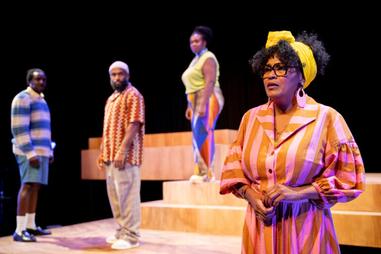 Vier mensen staan op het podium met een trappen attribuut, de oudste vrouw staat in het middelpunt met een ernstige uitdrukking.