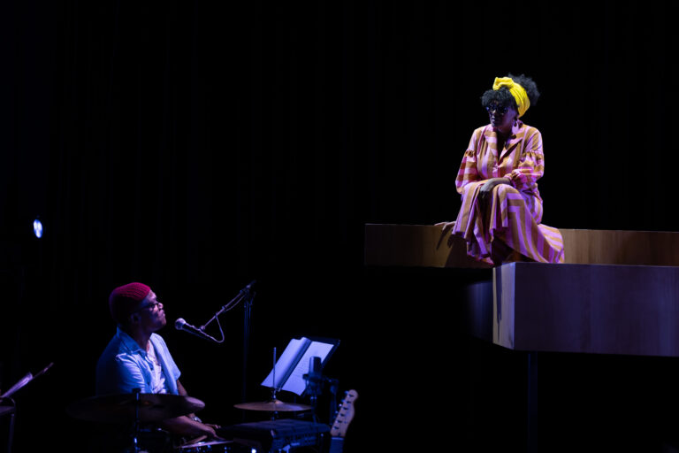 Een man achter een drumstel en een vrouw op een trap kijken naar elkaar terwijl ze verlicht worden.