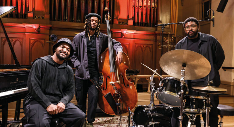 Drie jazzmuzikanten poseren met hun instrumenten in een concertzaal, met een groot pijporgel op de achtergrond.