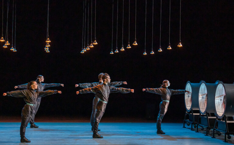 Groep kunstschaatsers in een rij op een verlichte ijsbaan, armen gestrekt langs de zijden.