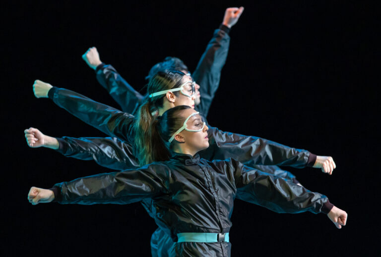 Danser in een zwart kostuum met wijd uitgestrekte armen tegen een donker podium.