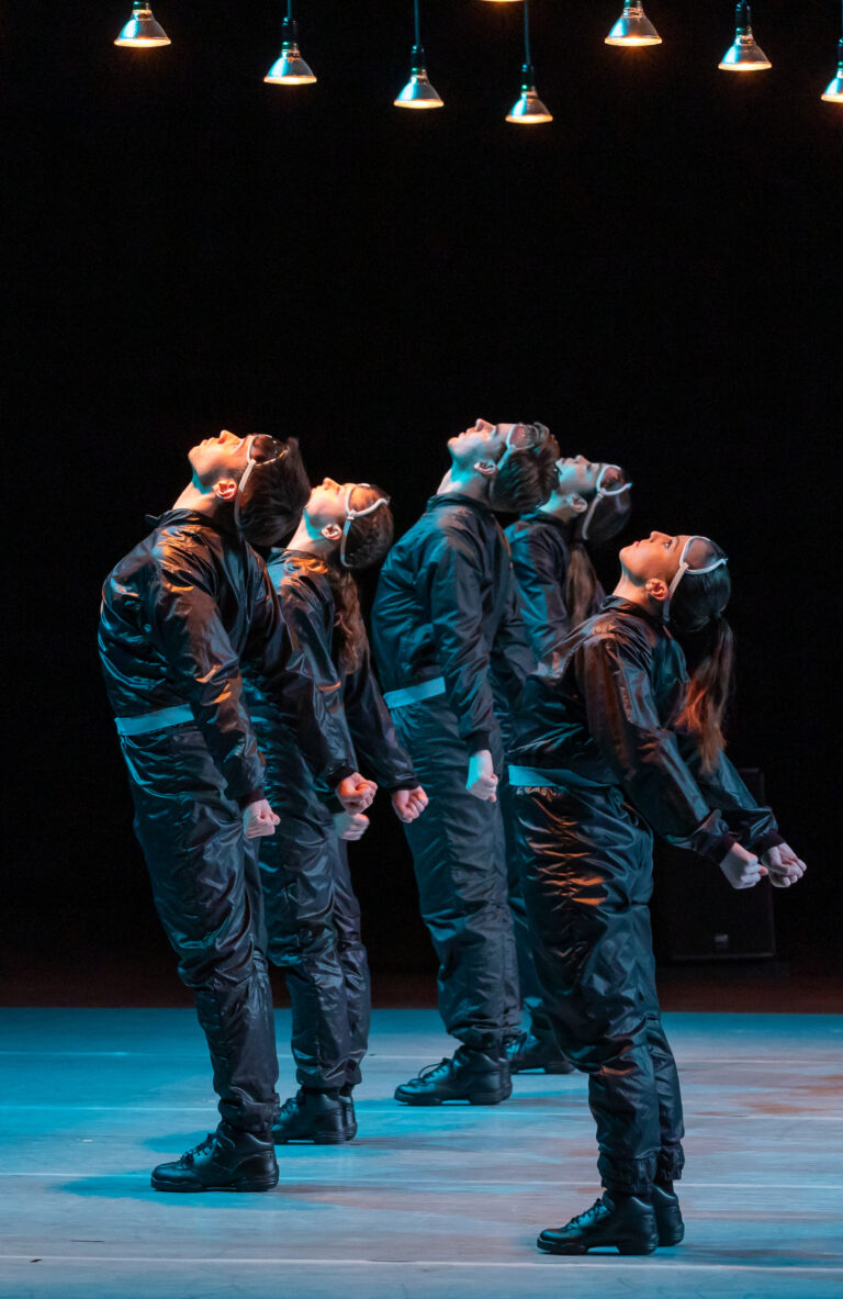 Vijf mensen in donkere kleding staan in een rij op een donker podium met blauw licht op de achtergrond.