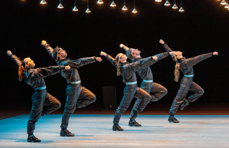 Een groep dansers in gelijkaardige denim outfits voert een choreografie uit op een podium.