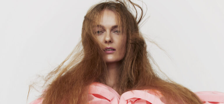 Een model draagt een avant-gardistische outfit met een roze bovenstuk met uitgesneden cirkels en oranje broek, met haar loshangend haar dat wild om haar hoofd valt.