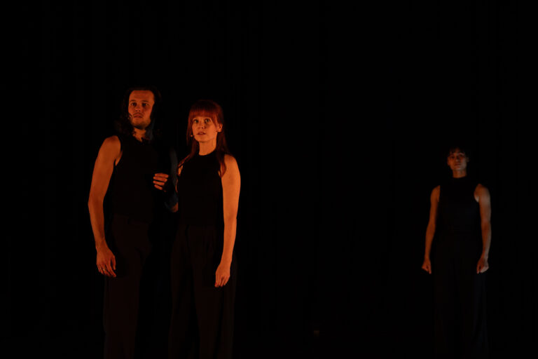 Twee personen, een man en een vrouw, staan dichtbij elkaar in donkere kleding op een schaars verlicht toneel, terwijl een derde persoon op de achtergrond staat, deels in de schaduw