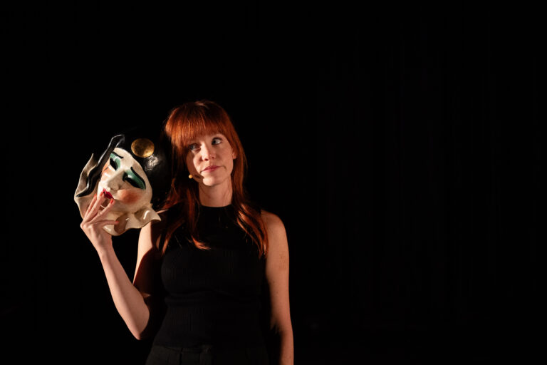 Een vrouw met rood haar, gekleed in een zwarte outfit, houdt een expressief beschilderd masker vast tegen haar hoofd. Het masker heeft een wit gezicht, donkere ogen en lippen, en een zwart hoofddeksel met een gouden cirkel. Ze kijkt omhoog met een licht bedenkelijke blik, terwijl ze op een donker toneel staat.