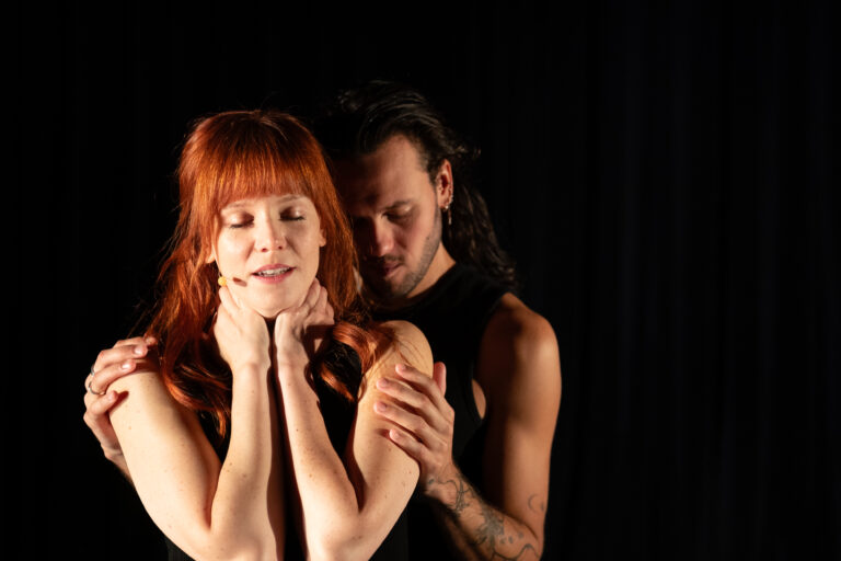 Een man en vrouw staan dichtbij elkaar, waarbij de man de vrouw vasthoudt en haar van achteren omarmt.