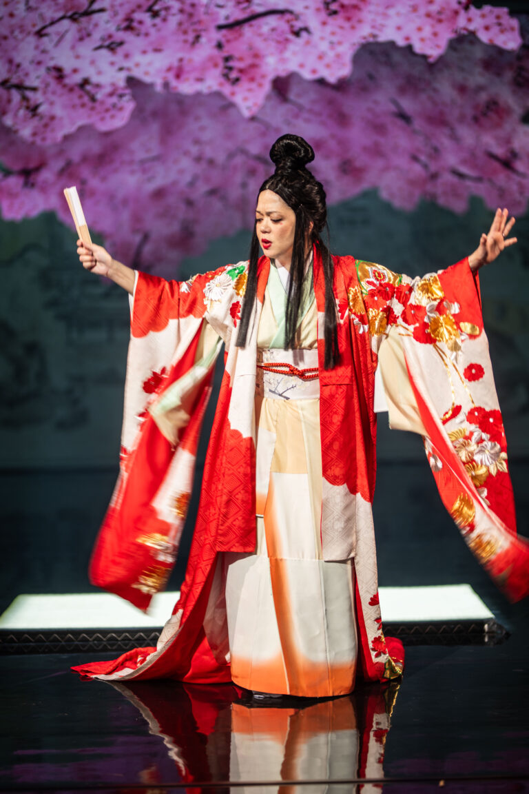 Een persoon in een rode en witte traditionele kimono danst op een podium met uitgestrekte armen tegen een roze bloemige achtergrond.