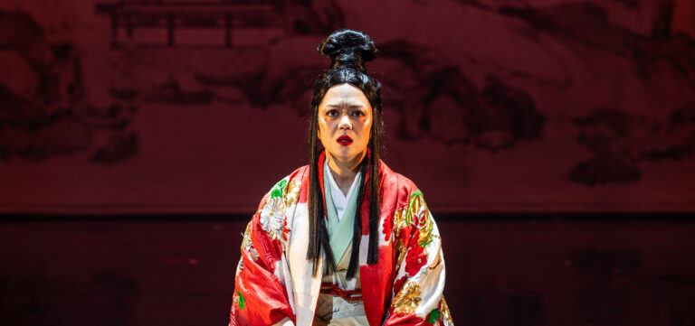 Een Chinese opera-artiest in een rood kostuum met bloemmotieven en opvallende gezichtsmake-up, staand tegen een donkerrode achtergrond.