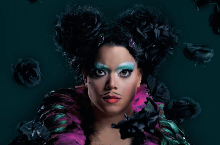 Actrice met donkere glitternde verenoutfit en een aanwezig makeup met een snorretje. Vanuit de lucht vallen zwarte bloemen.