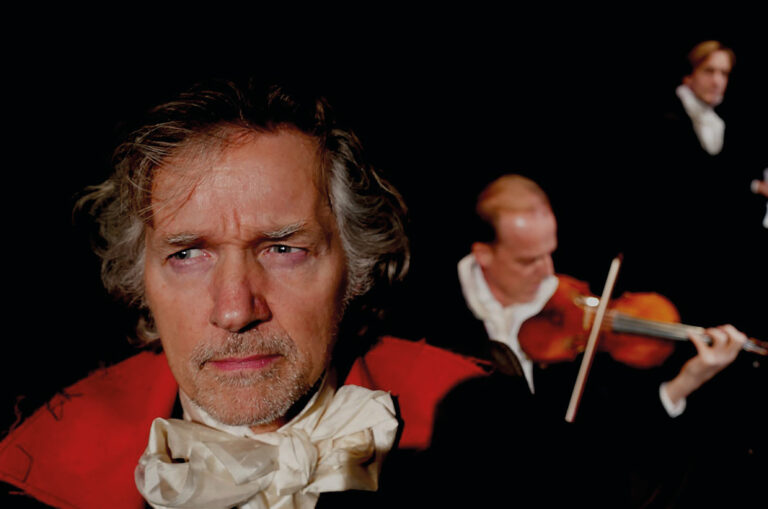 Thom Hoffman peinzend terwil er op de achtergrond een violist speelt